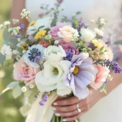 Une mariée tient une composition de bouquet de fleurs pastel, mêlant roses douces, anémones bleues et lavande, dans un écrin de verdure.