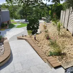 Un aménagement jardin gravier décoratif avec des pas japonais sur pelouse, des plantes et une clôture en bois.