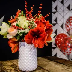 Magnifique composition amaryllis rouge et blanche, ornée de baies rouges, dans un vase texturé. Ballons festifs en arrière-plan.