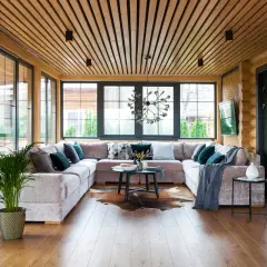 Aménagement véranda moderne avec grand canapé modulable, table basse, tapis peau de vache et plantes vertes. Lumière naturelle abondante.