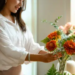 Une femme arrange une composition florale aux couleurs vives, mêlant feuillage et fleurs orangées.