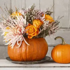 Une composition automnale avec des fleurs roses et oranges dans une citrouille évidée, à côté d'une petite citrouille.
