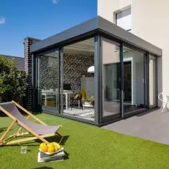 Aménagement véranda 15m2 moderne avec bureau et espace détente extérieur. Chaise longue, fruits et fauteuil blanc invitent au repos.