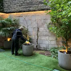 Un homme arrose un petit jardin moderne avec une pelouse artificielle et des plantes en pots.