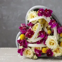 Une idée composition florale avec orchidées pour la Saint-Valentin. Roses crème, orchidées fuchsia et petites boules jaunes dans un contenant en forme de cœur.