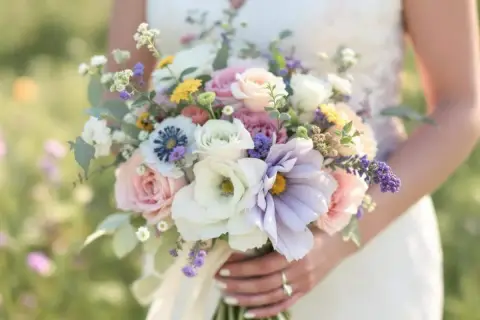 Une mariée tient une composition de bouquet de fleurs pastel, mêlant roses douces, anémones bleues et lavande, dans un écrin de verdure.
