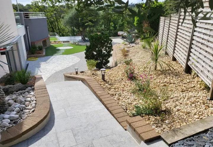 Un aménagement jardin gravier décoratif avec des pas japonais sur pelouse, des plantes et une clôture en bois.