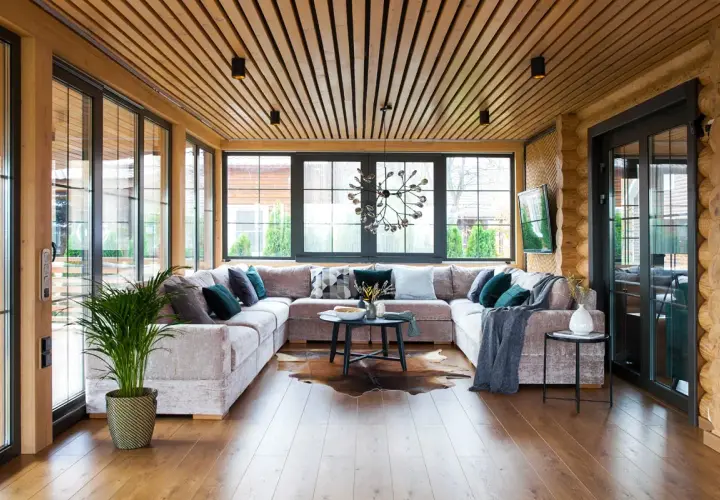 Aménagement véranda moderne avec grand canapé modulable, table basse, tapis peau de vache et plantes vertes. Lumière naturelle abondante.