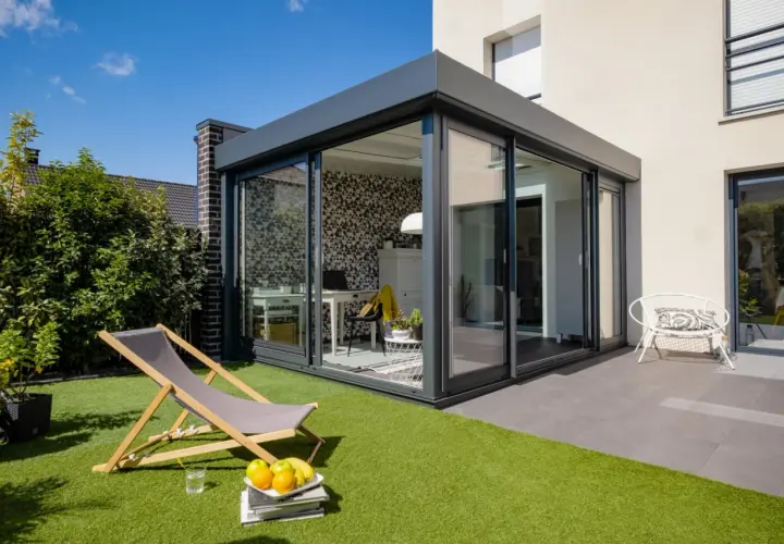 Aménagement véranda 15m2 moderne avec bureau et espace détente extérieur. Chaise longue, fruits et fauteuil blanc invitent au repos.