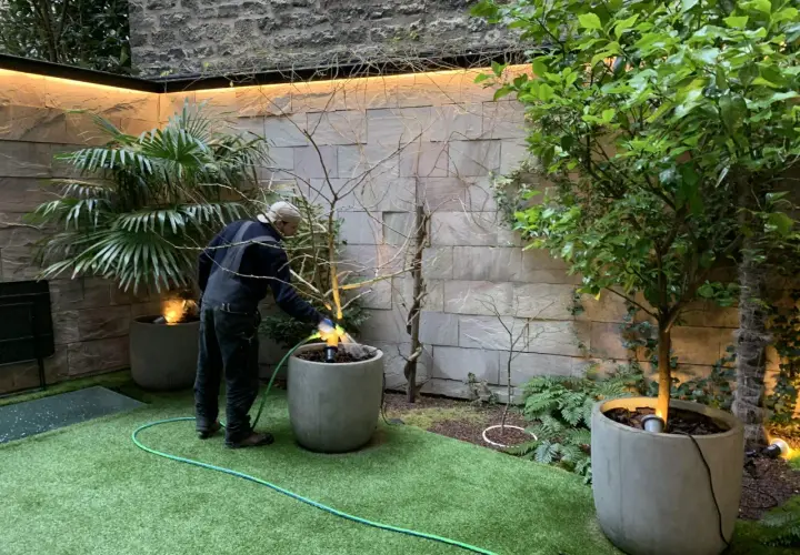 Un homme arrose un petit jardin moderne avec une pelouse artificielle et des plantes en pots.