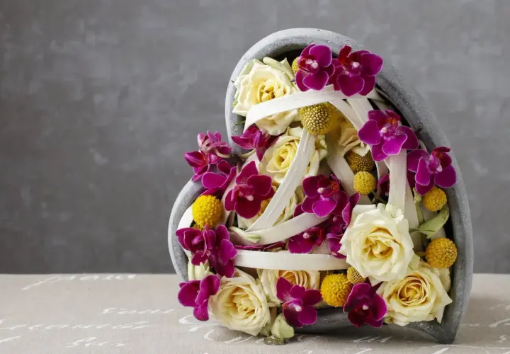 Une idée composition florale avec orchidées pour la Saint-Valentin. Roses crème, orchidées fuchsia et petites boules jaunes dans un contenant en forme de cœur.