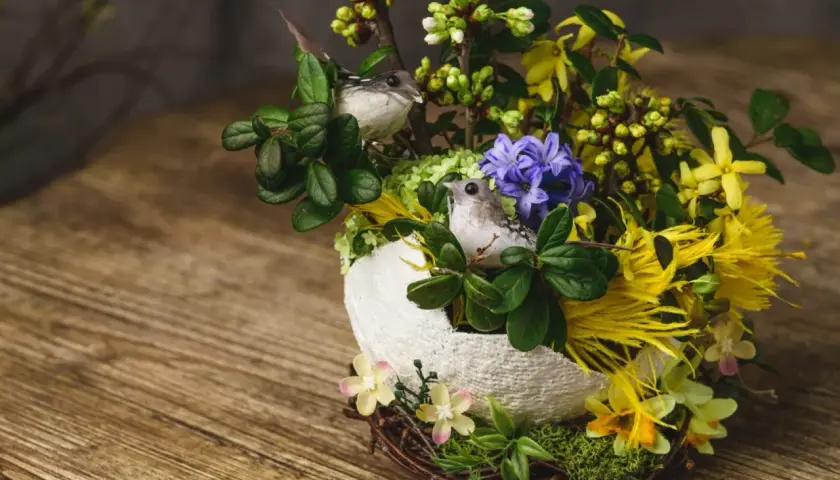 Un nid de paques composition florale de paques avec deux petits oiseaux, des fleurs jaunes et violettes, et des branches vertes sur un fond en bois.