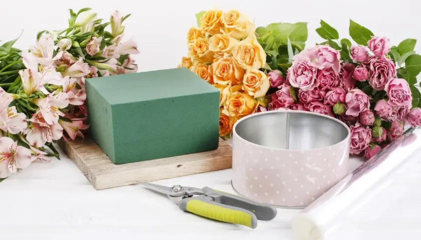 Préparation d'une petite composition florale simple : roses roses et jaunes, alstroemérias, mousse florale, boîte, ciseaux et film.