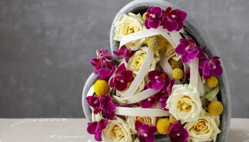 Une idée composition florale avec orchidées pour la Saint-Valentin. Roses crème, orchidées fuchsia et petites boules jaunes dans un contenant en forme de cœur.