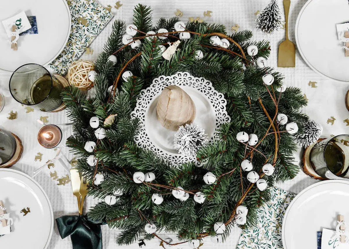 Un centre de table Noël nature avec une couronne de sapin, des clochettes blanches et des éléments décoratifs rustiques.