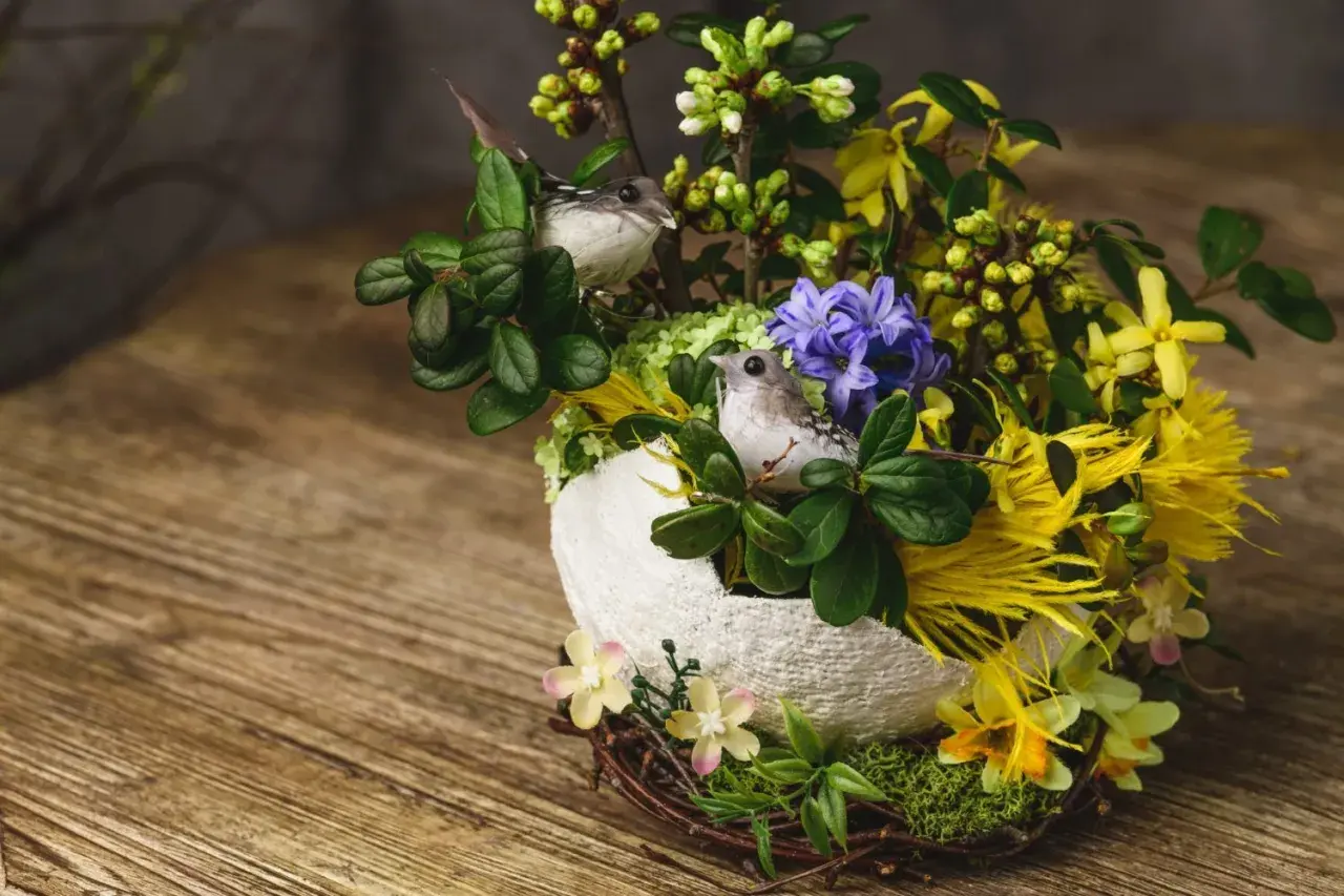 Un nid de paques composition florale de paques avec deux petits oiseaux, des fleurs jaunes et violettes, et des branches vertes sur un fond en bois.