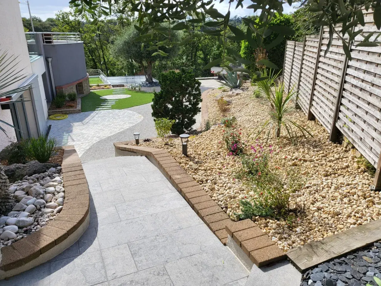 Un aménagement jardin gravier décoratif avec des pas japonais sur pelouse, des plantes et une clôture en bois.