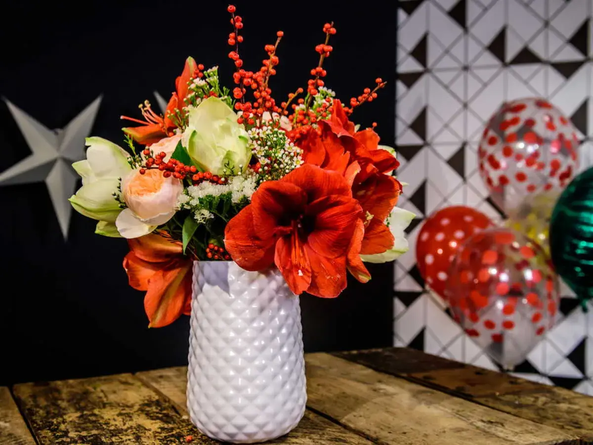 Magnifique composition amaryllis rouge et blanche, ornée de baies rouges, dans un vase texturé. Ballons festifs en arrière-plan.