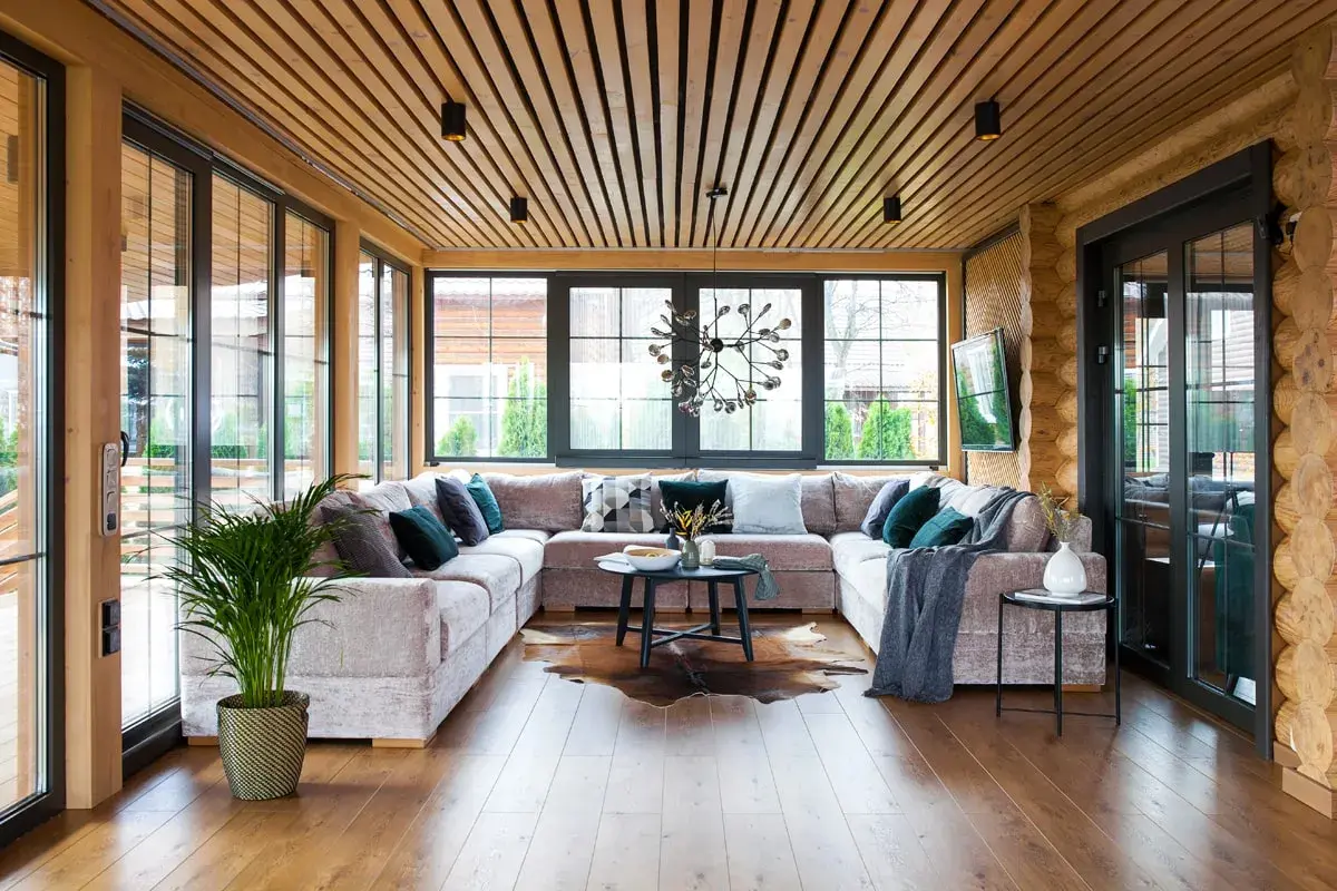 Aménagement véranda moderne avec grand canapé modulable, table basse, tapis peau de vache et plantes vertes. Lumière naturelle abondante.