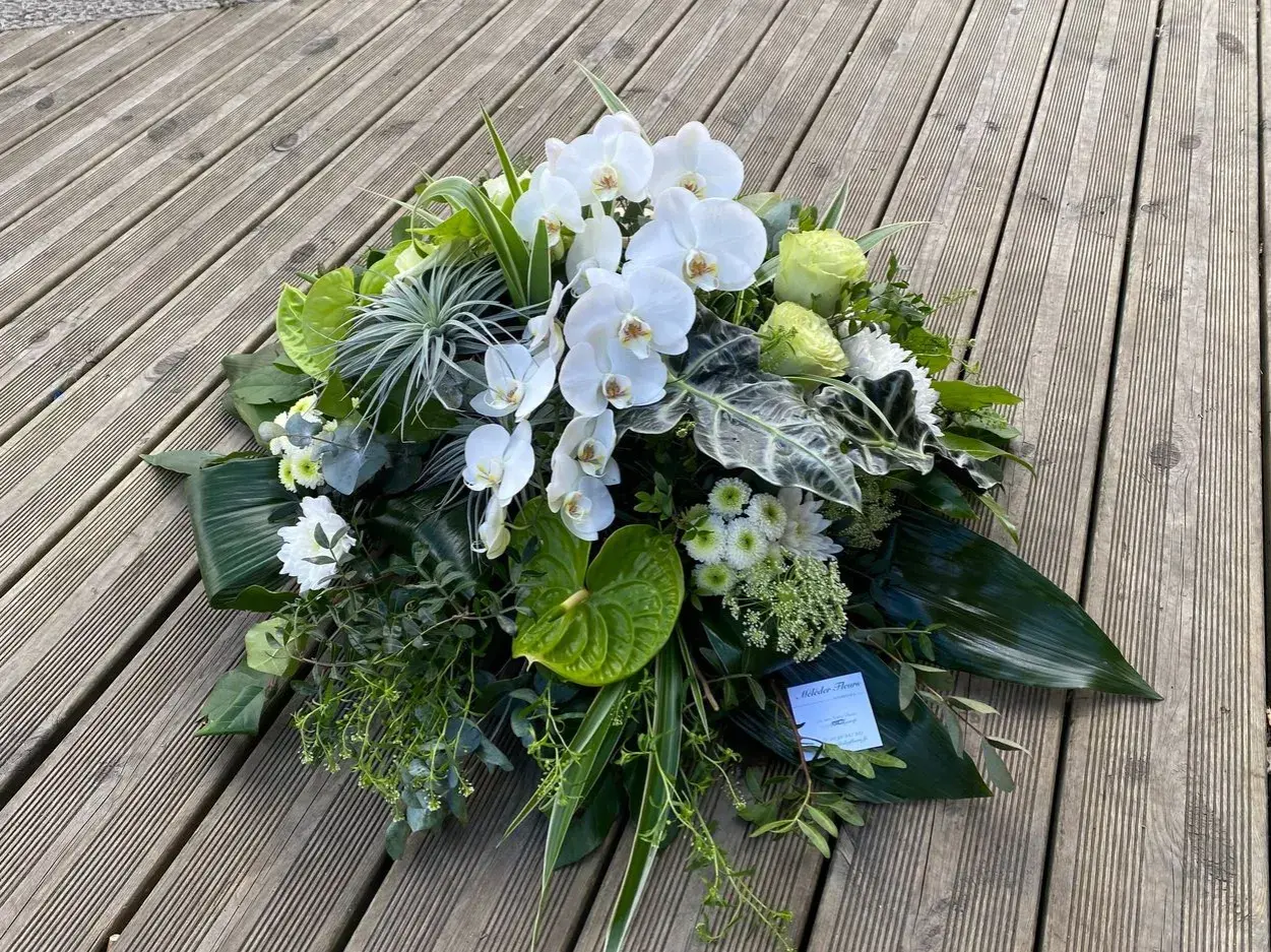 Un arrangement floral blanc et vert, composé d'orchidées, de roses et d'anthuriums, évoque un coussin de deuil moderne.