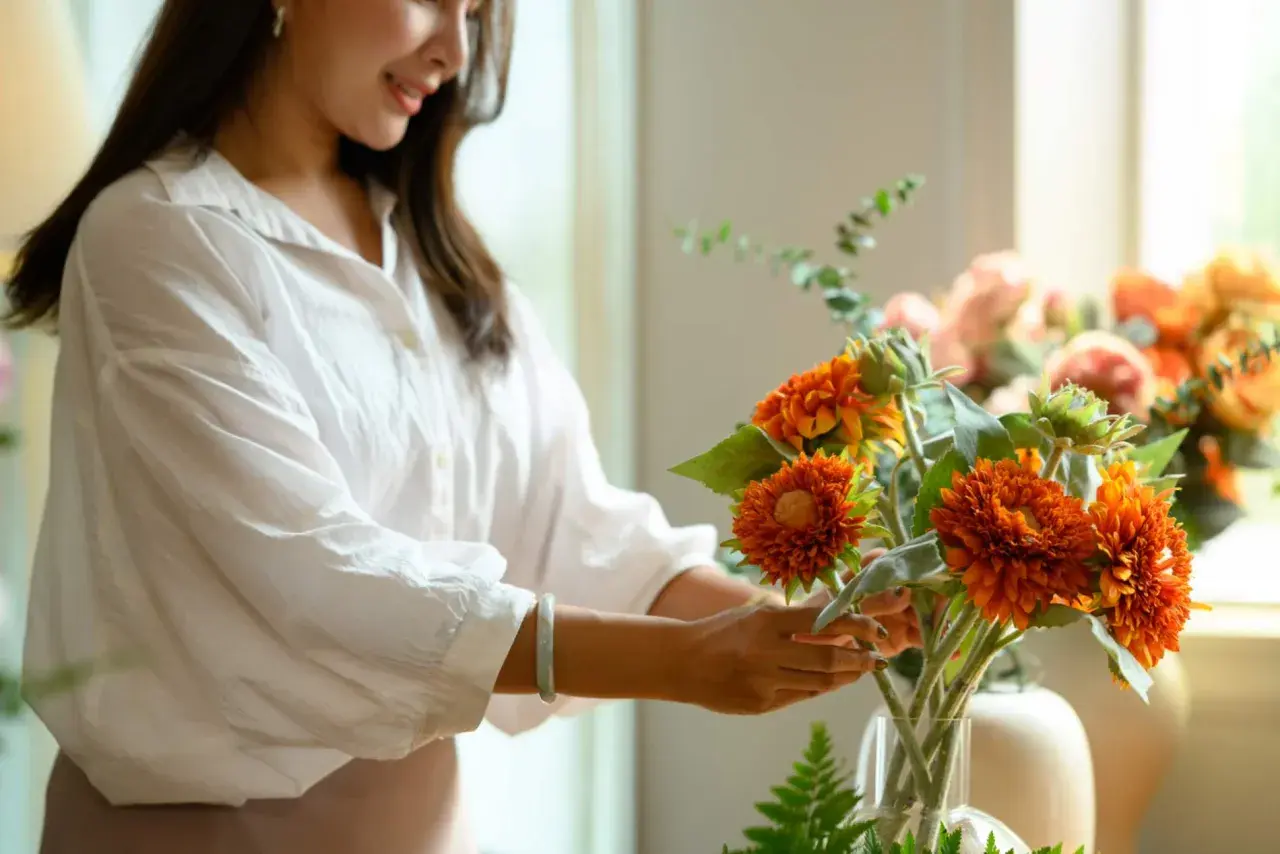 Une femme arrange une composition florale aux couleurs vives, mêlant feuillage et fleurs orangées.