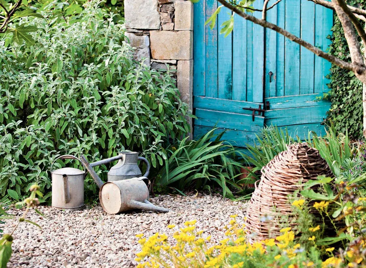 Un coin de jardin charmant avec des arrosoirs anciens, une porte bleue et des plantes luxuriantes, parfait pour créer un massif sans entretien.