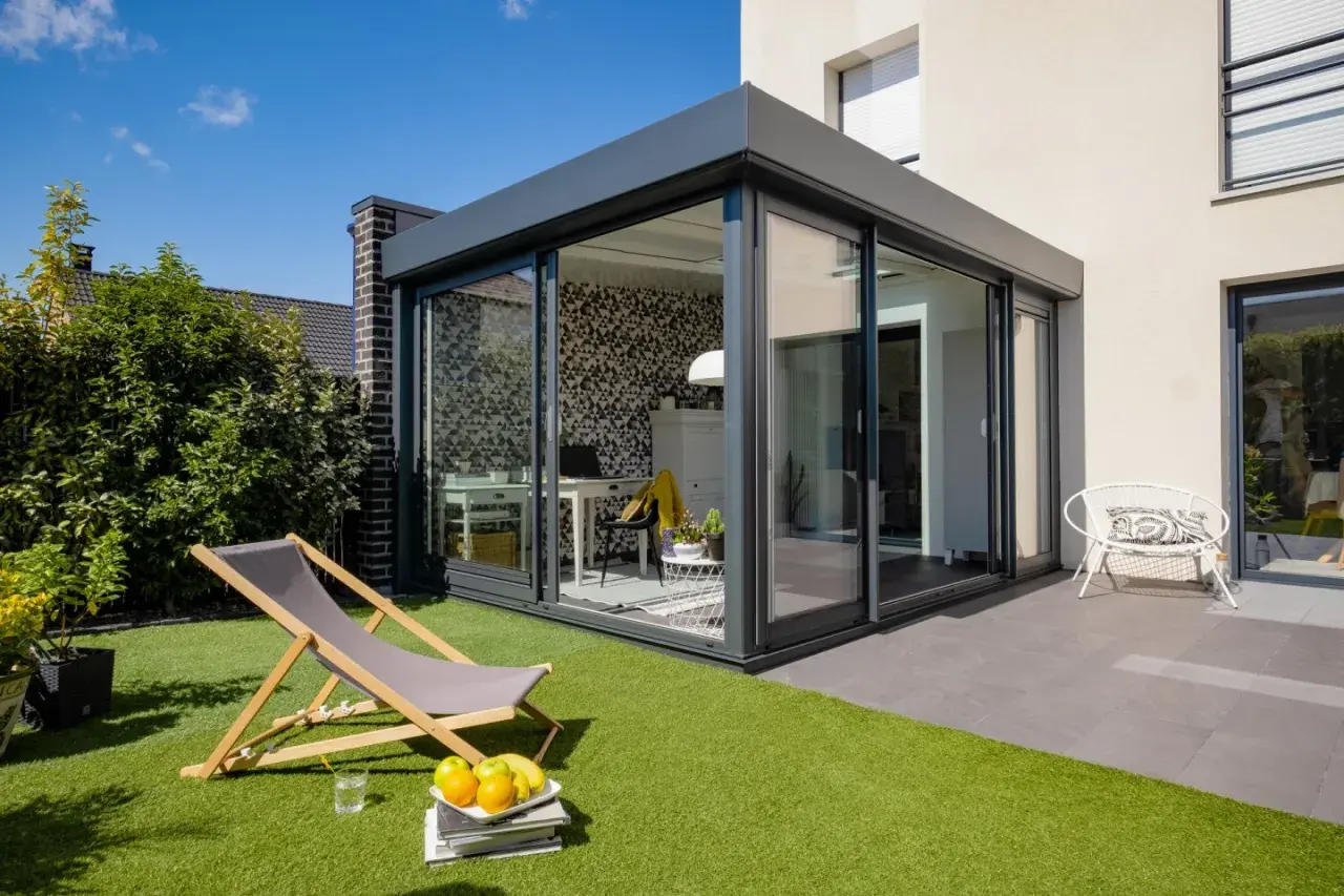 Aménagement véranda 15m2 moderne avec bureau et espace détente extérieur. Chaise longue, fruits et fauteuil blanc invitent au repos.