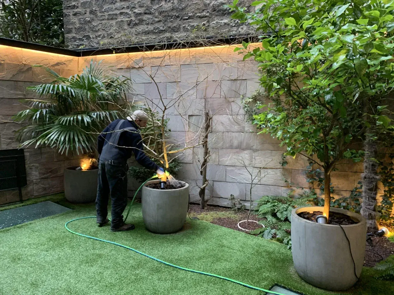 Un homme arrose un petit jardin moderne avec une pelouse artificielle et des plantes en pots.
