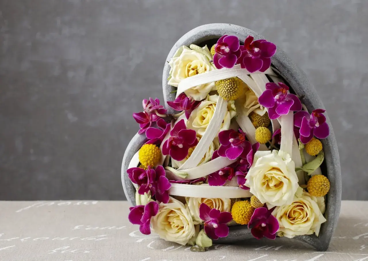 Une idée composition florale avec orchidées pour la Saint-Valentin. Roses crème, orchidées fuchsia et petites boules jaunes dans un contenant en forme de cœur.
