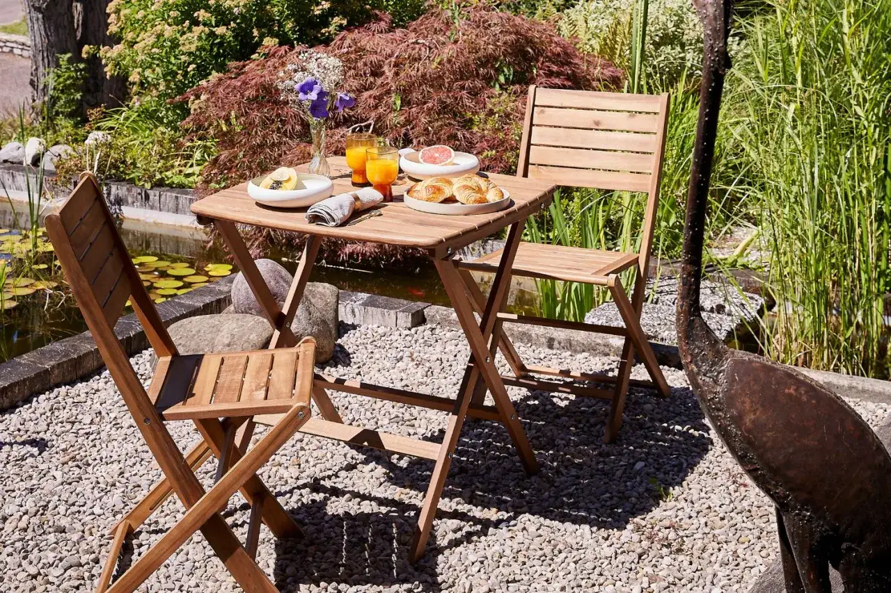 Petit d&eacute;jeuner sur une petite terrasse en bois, pr&egrave;s d'un &eacute;tang. Deux chaises pliantes et une table avec croissants, jus d'orange et fruits.