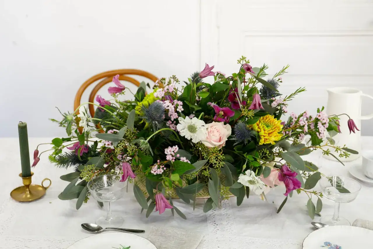 Un magnifique bouquet de fleurs sauvages, aux teintes roses, violettes et jaunes, est pos&eacute; sur une table dress&eacute;e. La mousse pour piquer des fleurs sert de base &agrave; cette composition florale.
