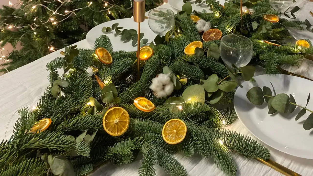 Un magnifique centre de table No&euml;l nature avec des branches de sapin, des rondelles d'orange s&eacute;ch&eacute;e, des pommes de pin et des guirlandes lumineuses.