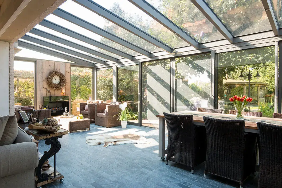 Am&eacute;nagement v&eacute;randa lumineux avec salon confortable, chemin&eacute;e et salle &agrave; manger. Vue sur jardin verdoyant.