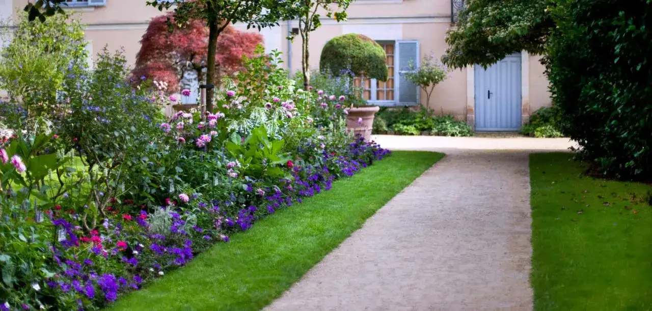 Un chemin de gravier serpente à travers un jardin luxuriant, bordé d'un parterre de fleurs éclatant de roses, de violettes et de touches de rose.
