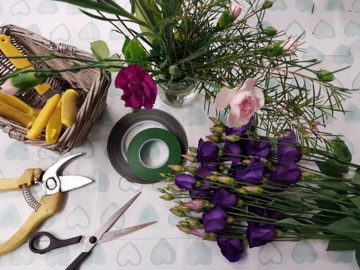 Matériel pour une composition florale à faire soi-même : fleurs violettes, roses, roses foncées, ciseaux, sécateur, ruban floral et panier.