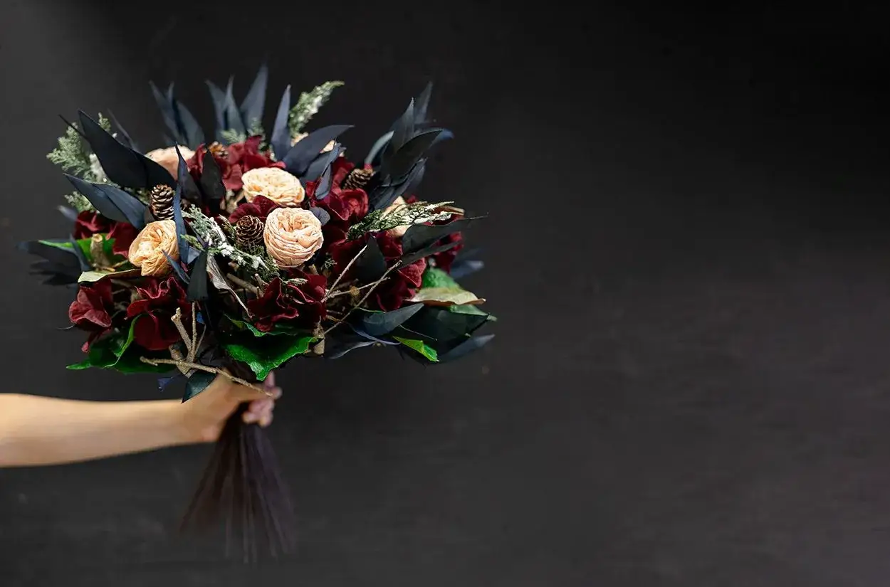 Une main tient une composition de bouquet de fleurs aux tons sombres, avec des roses pâles, des fleurs bordeaux et des feuillages bleutés.