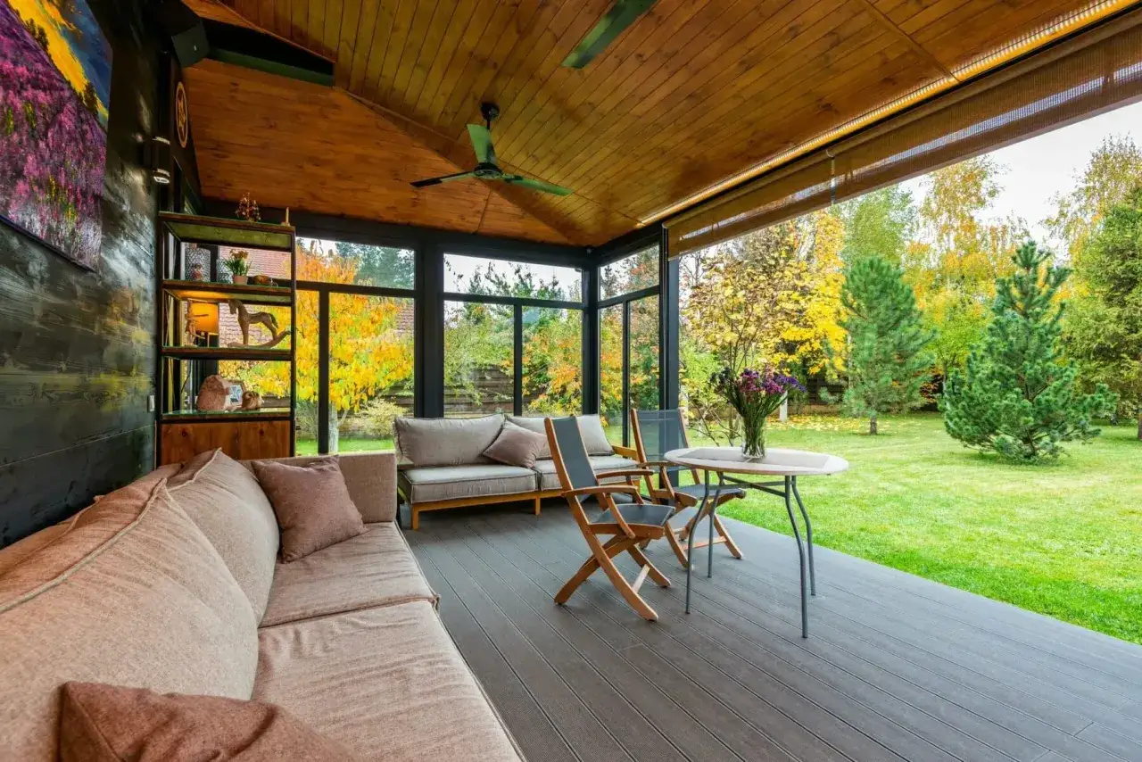 Am&eacute;nagement v&eacute;randa 15m2 avec canap&eacute;s confortables, table et chaises, offrant une vue imprenable sur un jardin verdoyant en automne.