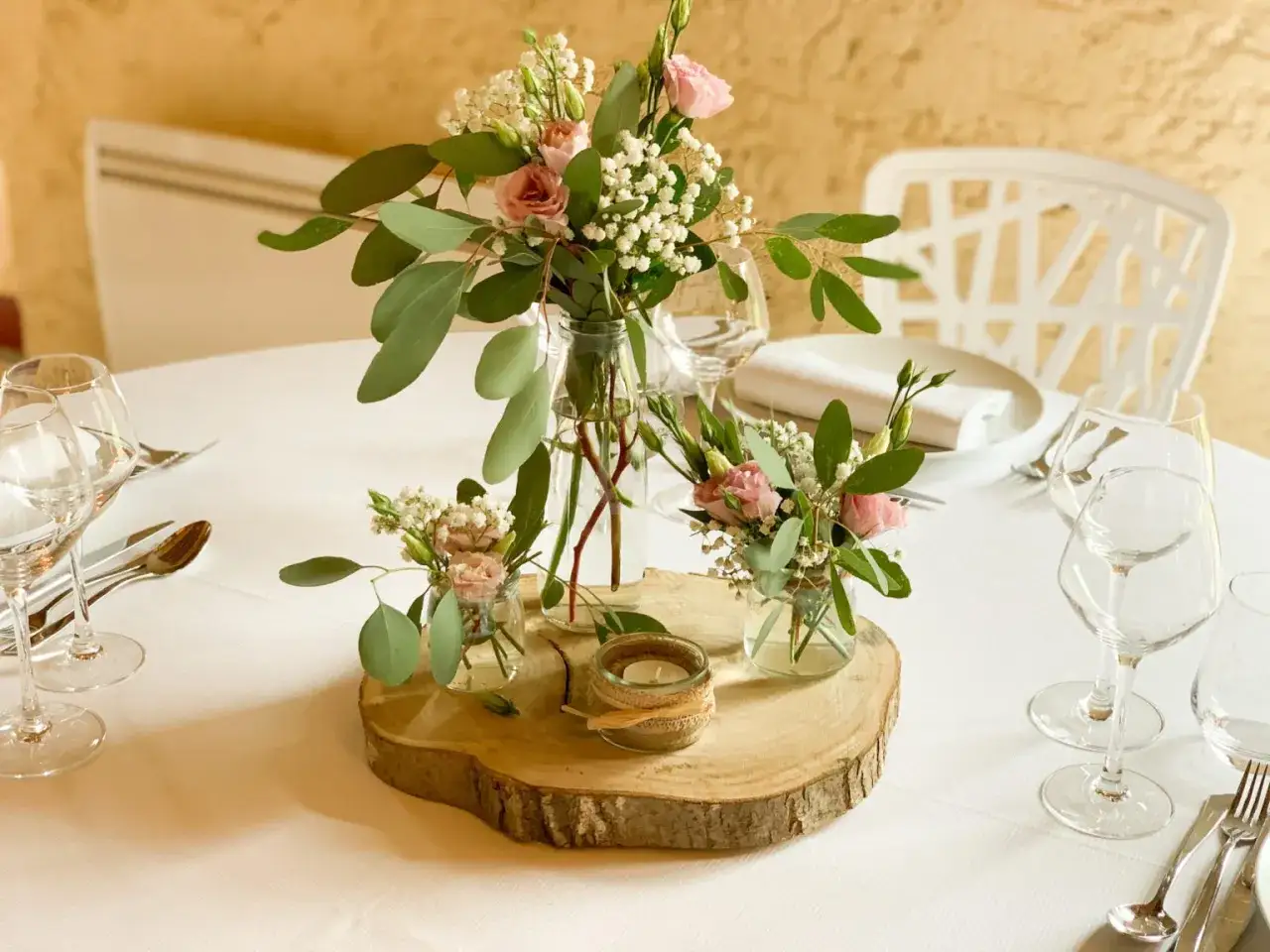 D&eacute;coration champ&ecirc;tre pour un mariage : des fleurs d&eacute;licates dans des vases en verre sur une tranche de bois, entour&eacute;es de bougies et de vaisselle &eacute;l&eacute;gante.